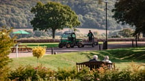 Two guests sitting on a bench in the fore ground and watching the grounds keepers driving by in a maintenance vehicle, surrounded by beautiful greenery