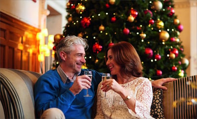 A man and woman toast with champagne on a couch beside a beautifully decorated Christmas tree, celebrating the festive season.