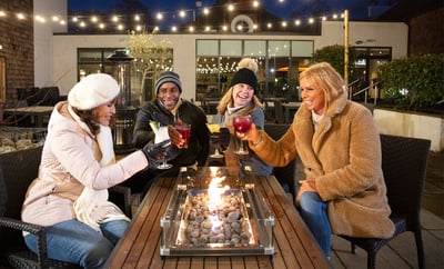 Four friends joyfully toasting drinks over a festive fire pit, creating a warm and inviting atmosphere.